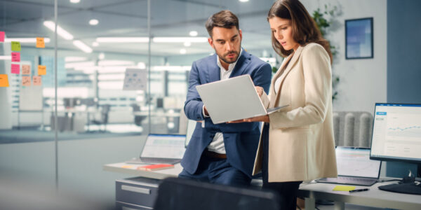 Young Manager Talking with Experienced Colleague while Using Laptop Computer in Office. Colleagues Discuss Business, Financial and Marketing Projects. Specialists Work in Diverse Team.